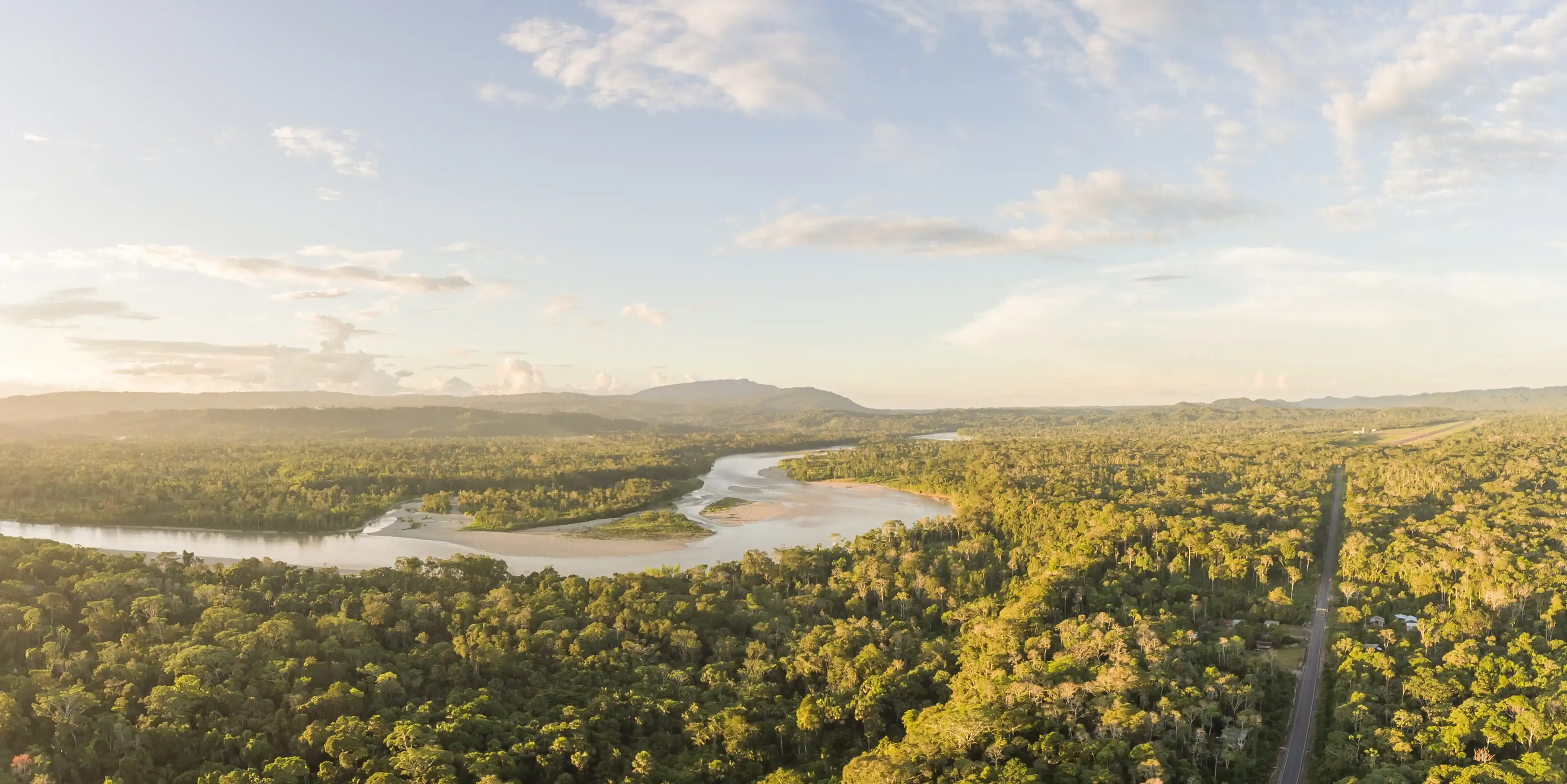 Aerial panorama of an Amazonian highway in Ecuador at dusk with Rio Napo and Galeras mountain in the background. Roads bring colonization and destruction of the rainforest to the Amazon Basin.