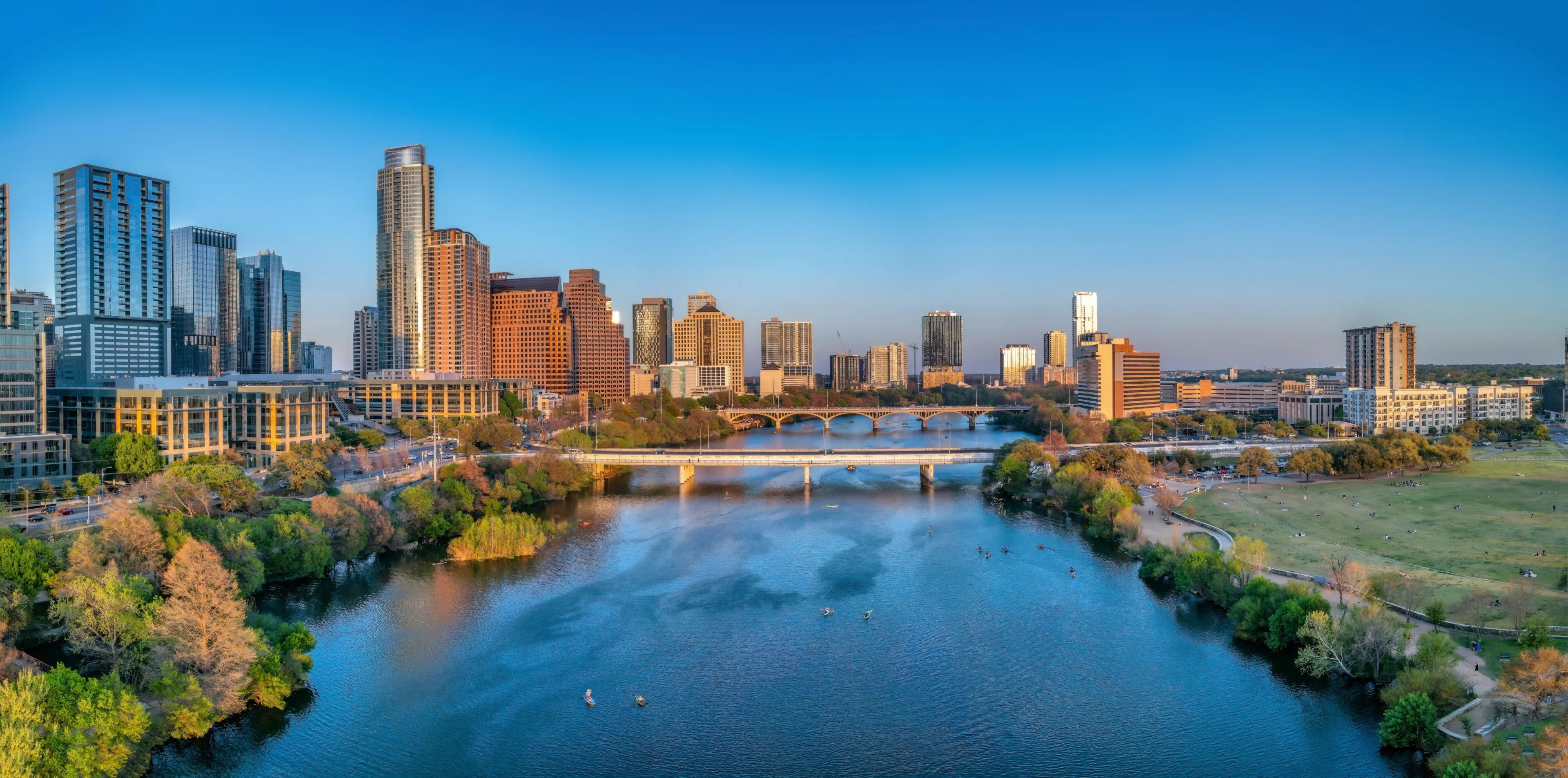 Colorado River near the district urban area of Austin, Texas. There are bridges over the river and a view of skyscraper buildings on the left and large field at the right side against the sunset sky.