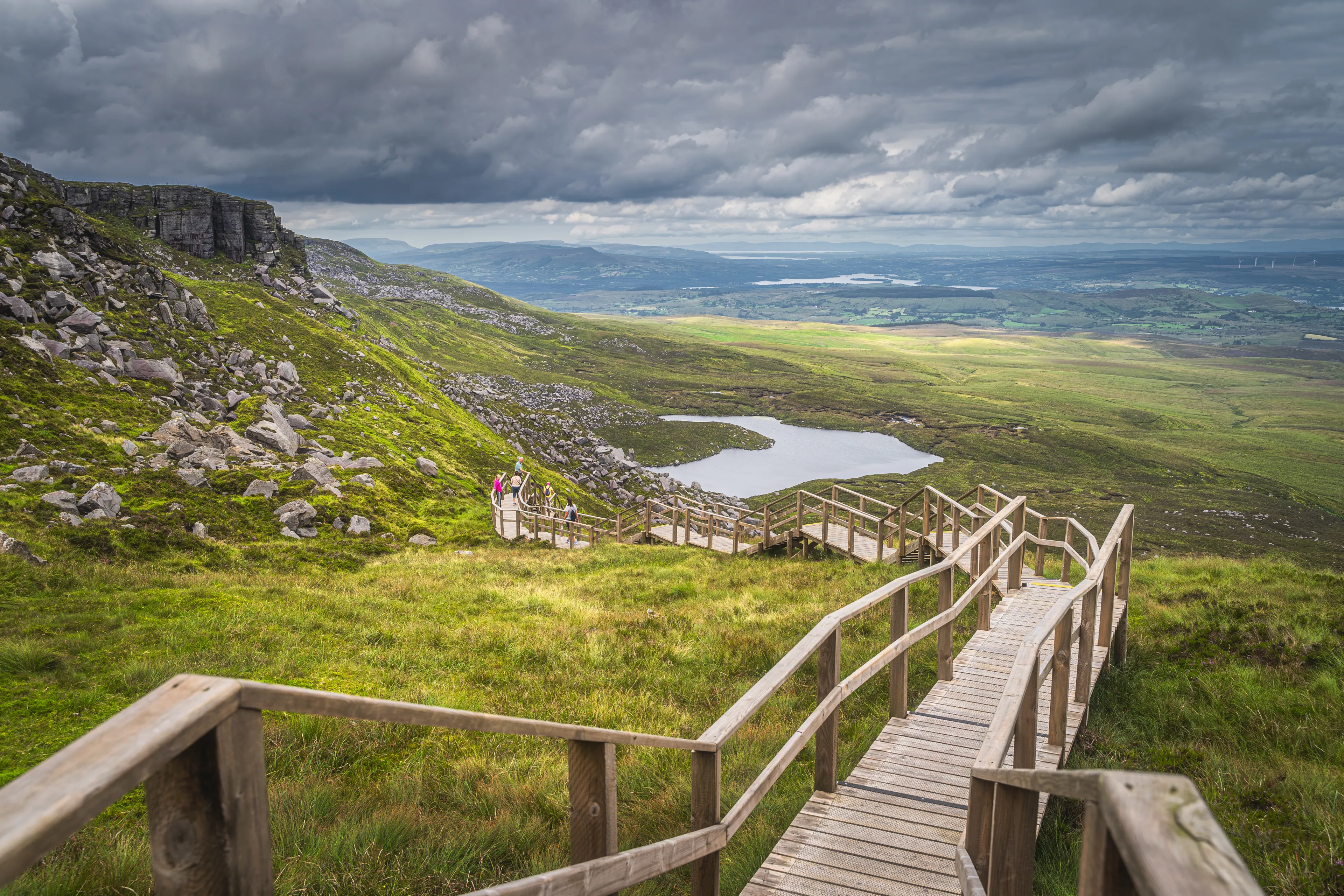 People enjoying a walk on steep stairs of wooden boardwalk in Cuilcagh Mountain Park with a view on lake and valley below, Northern Ireland