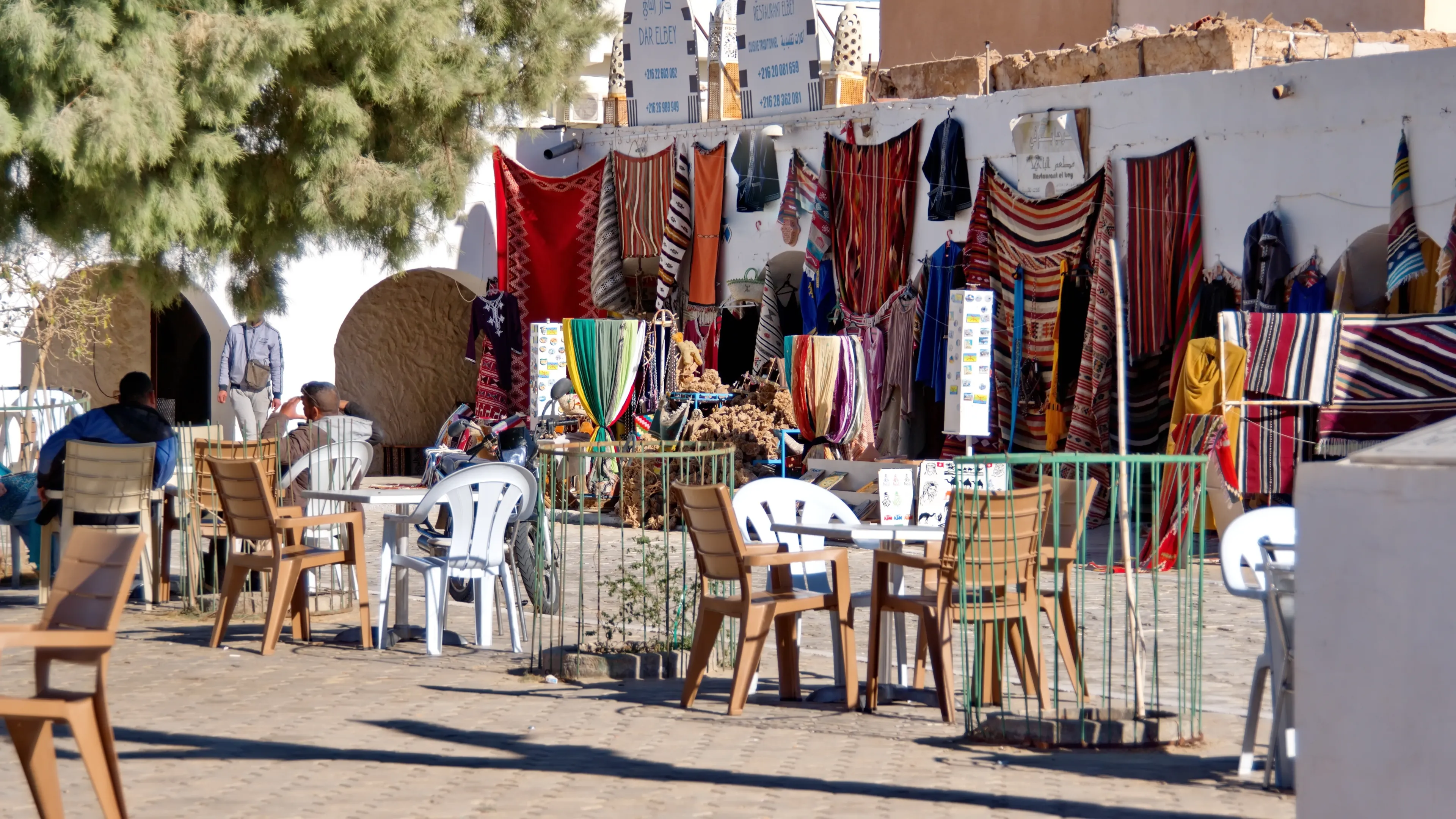 Douz, Tunisia - March 5, 2023: Cafe, with tables and chairs in the courtyard, set up in front of a stall selling colorful textiles in the market
