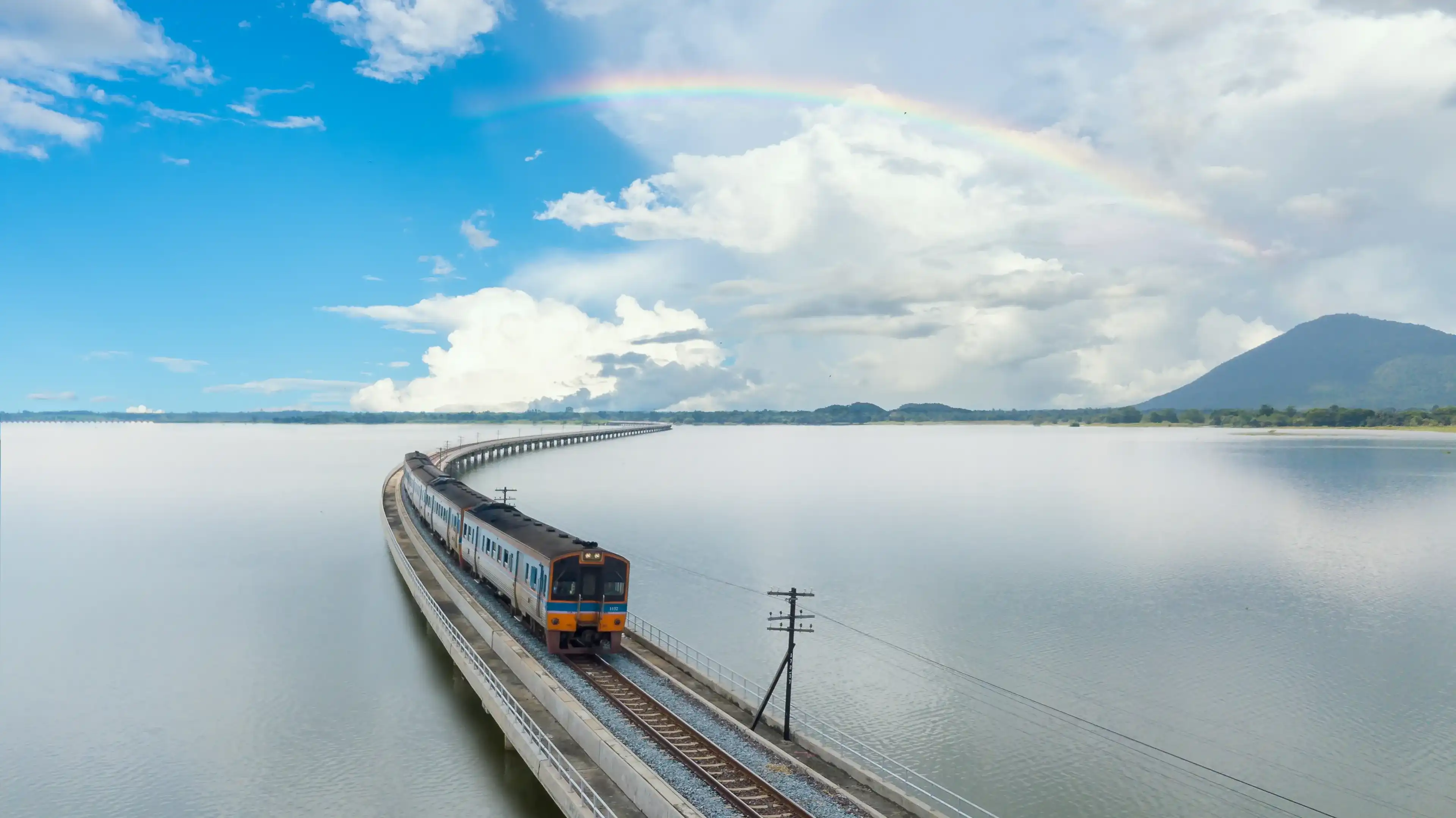 Aerial view of Travel train at speed on a floating railway bridge above the lake of Pa Sak Jolasid dam with the rainbow is background at Lopburi, unseen Thailand. Amazing floating train in Thailand