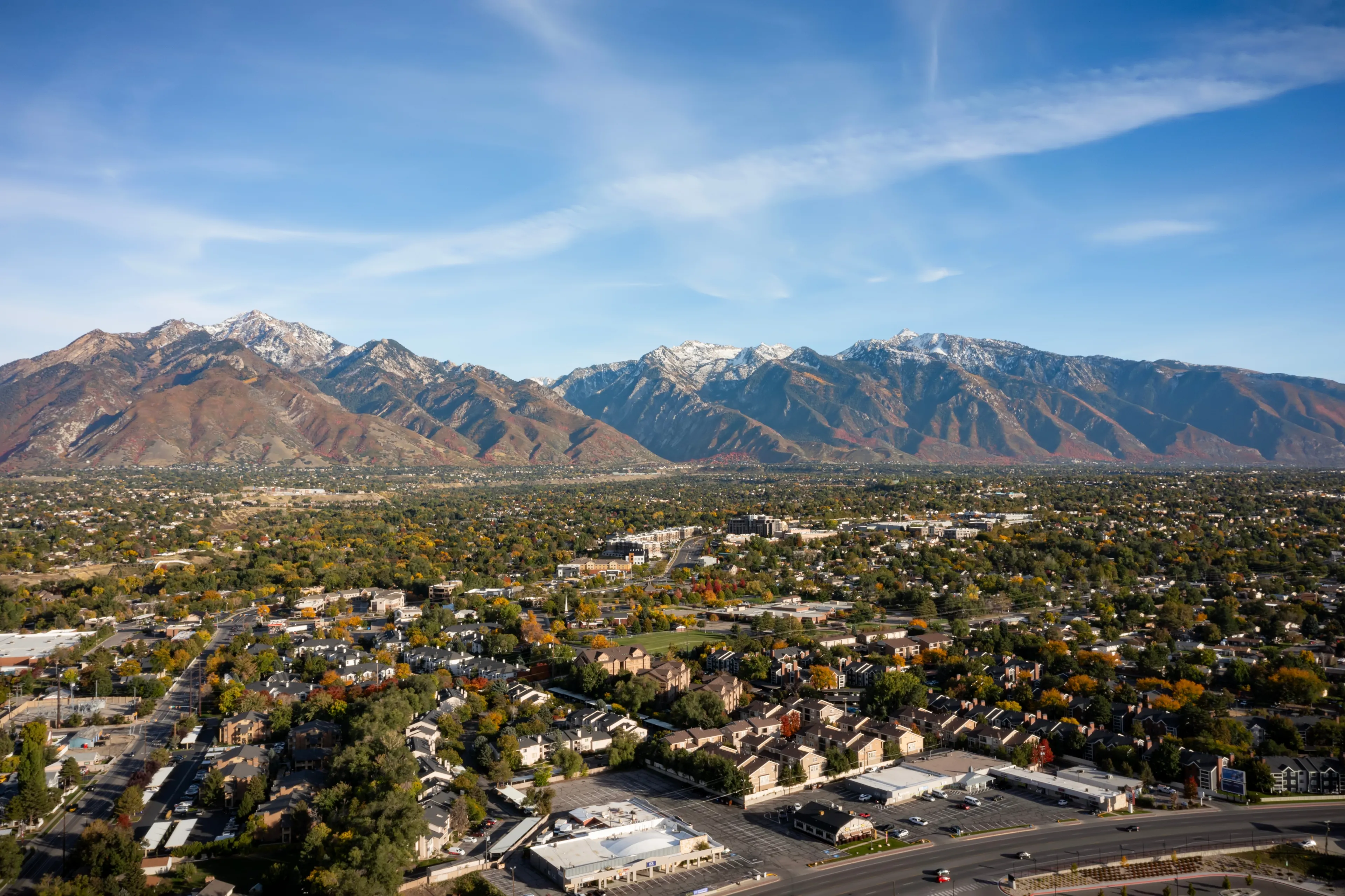 Aerial drone wide landscape shot of the stunning snowcapped rocky mountains of Utah with Salt Lake county below full of buildings and colorful trees on a warm sunny fall day