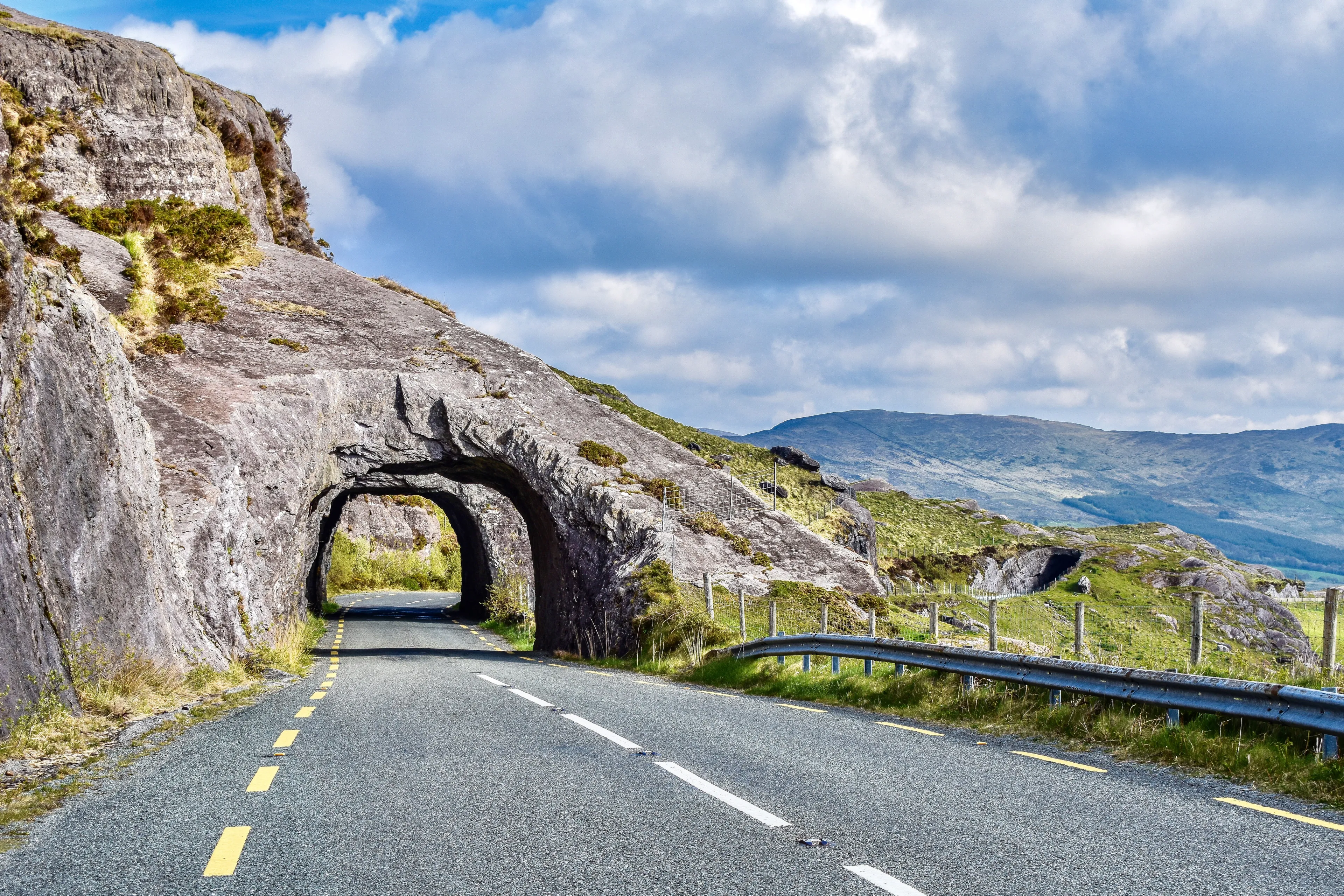 Kenmare Road, County Kerry, Ireland, stone arch tunnels made from gray stone showing lined highway N71 with gray guardrail in the foreground backed by light green mountains and a mostly cloudy sky.