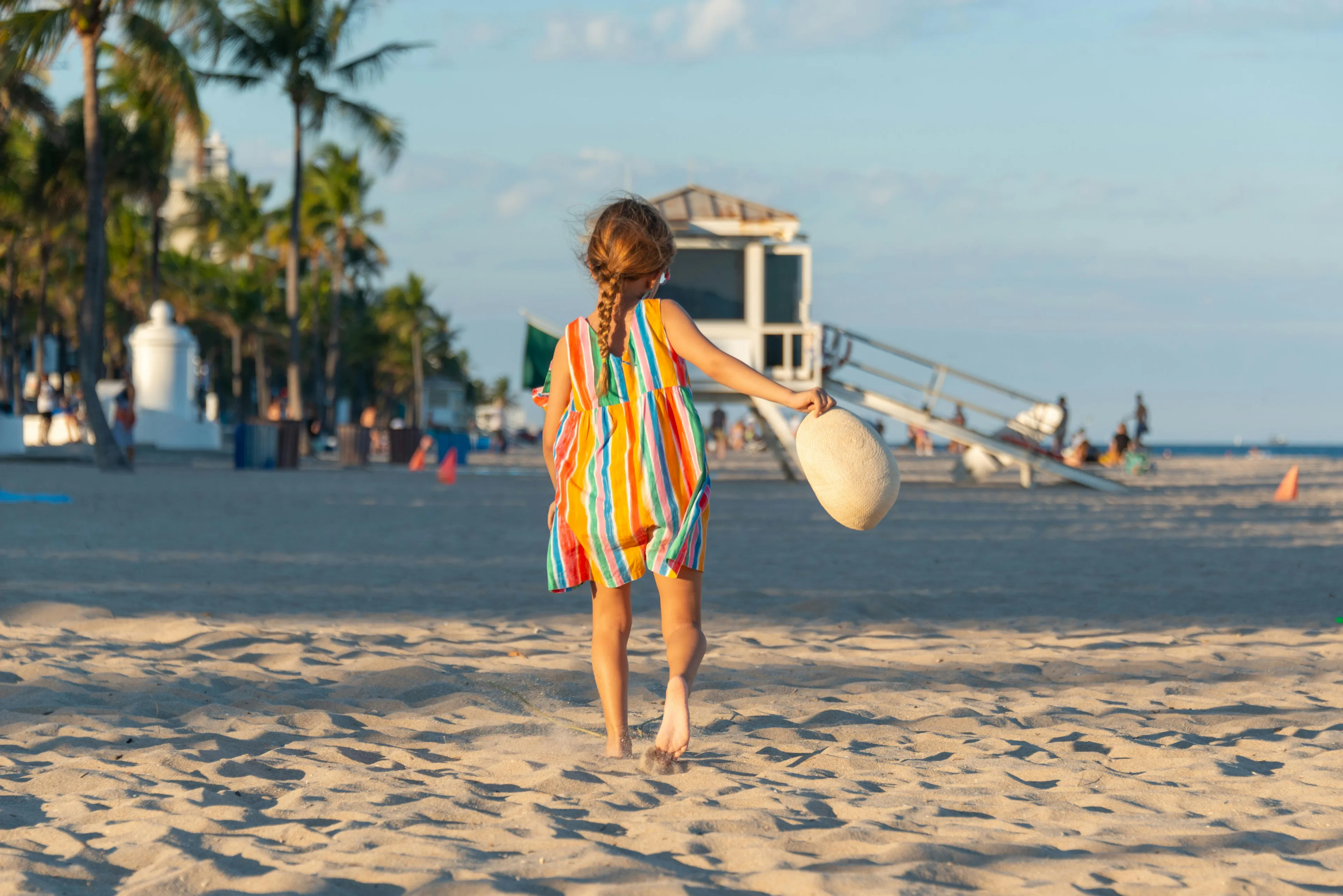 Young girl in colorful dress walking on beach, holding hat. Summer vacation vibe.