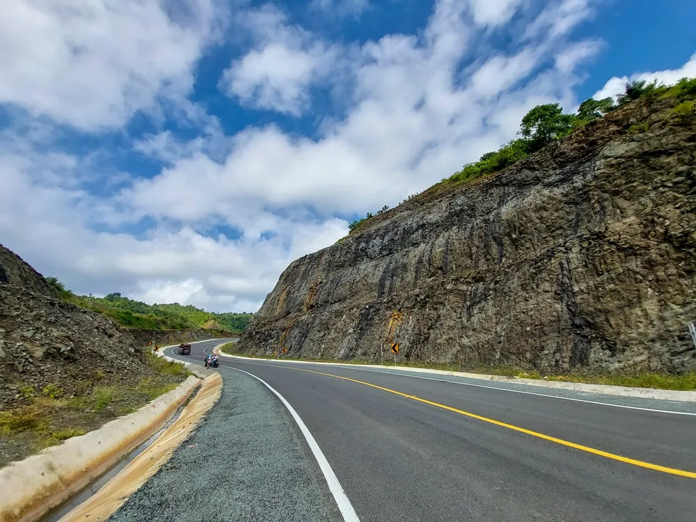 Scenic view of the Southern Cross Route in Tulungagung, Indonesia. A smooth winding asphalt road cutting through rocky hills under a bright blue sky with fluffy clouds.