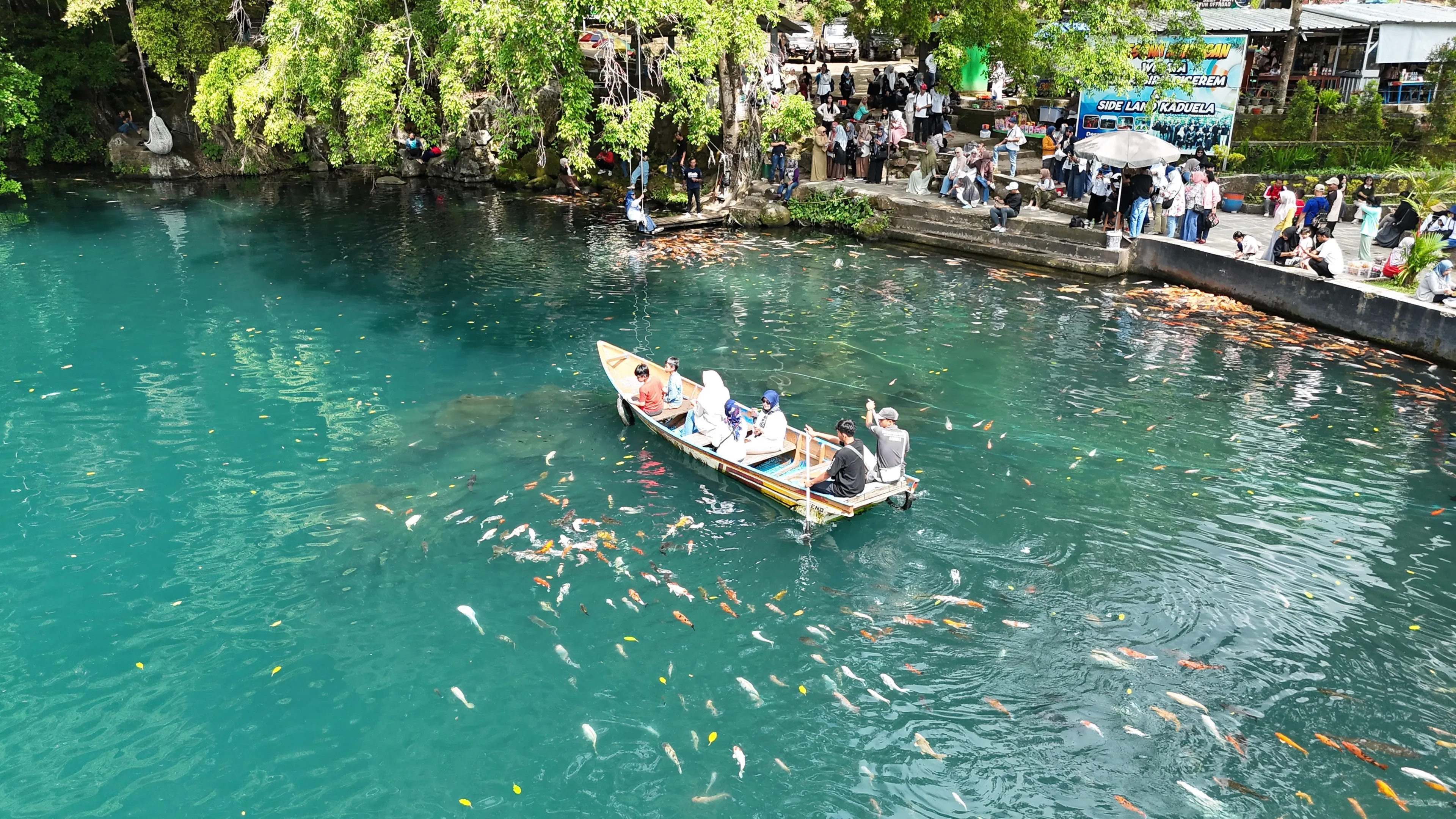 Kuningan - Indonesia. June 29, 2025. Aerial view of Telaga Biru Cicerem, West Java, with a tourist boat floating on crystal-clear blue spring water and people lining up to get photos