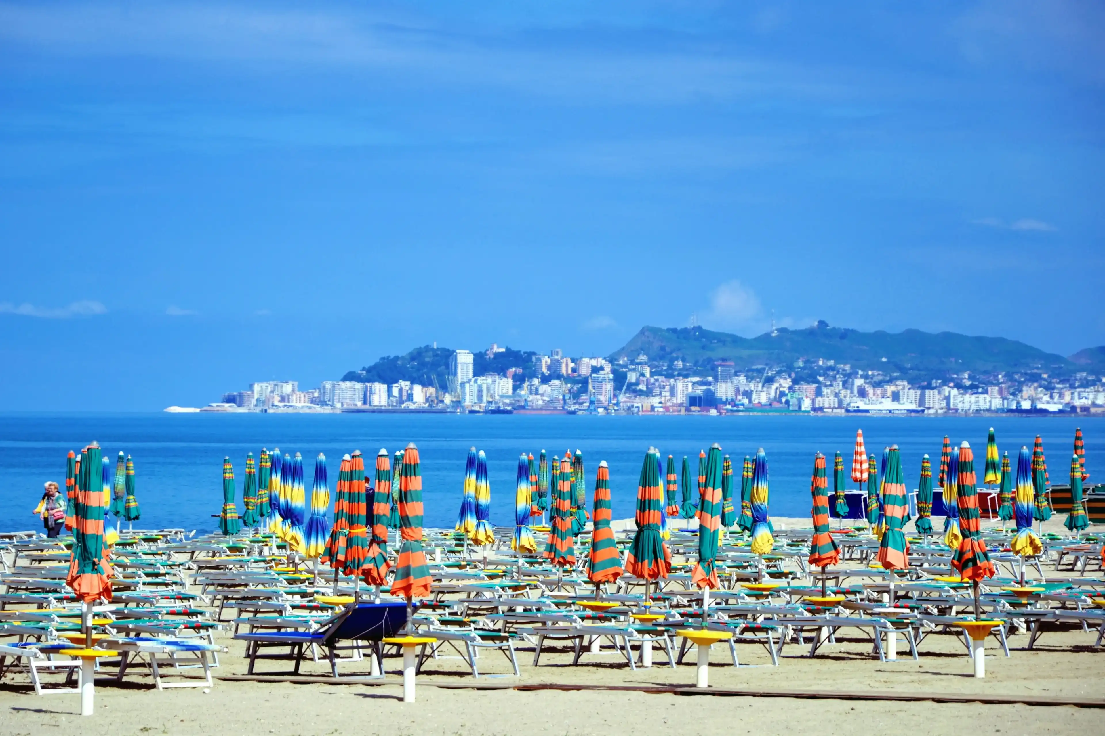 Closed umbrellas and empty chaise lounges at the sandy beach. Beach chairs, beds and umbrellas on the Albanian beach before summer holiday season. Golem, Durres, Albania. Durres city in a distance Closed umbrellas and empty chaise lounges at the sandy beach. Beach chairs, beds and umbrellas on the Albanian beach before summer holiday season. Golem, Durres, Albania. Durres city in a distance