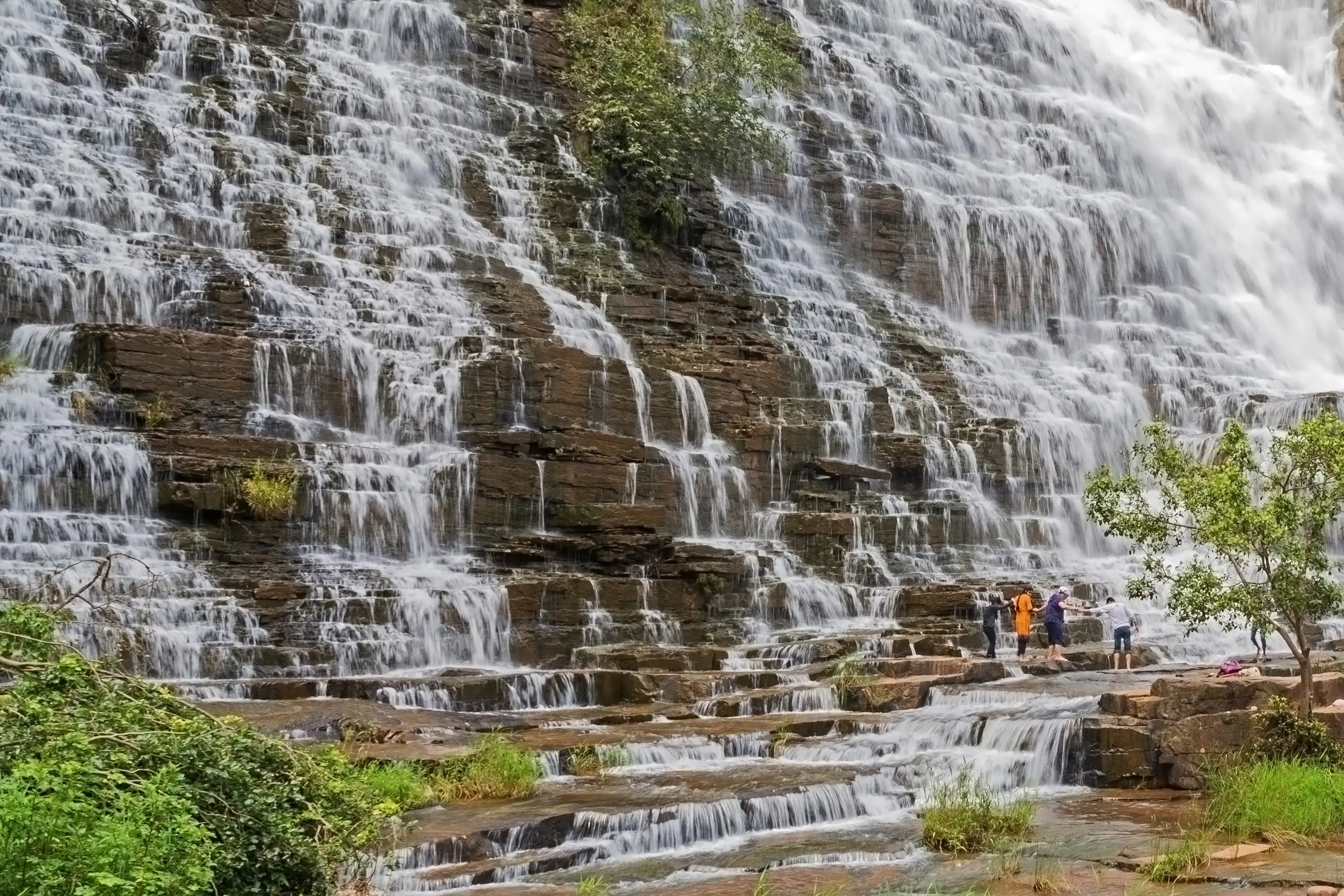 A group of friends enjoying the spectacular, magnificent sight of the milky waterfall cascaded down over the rocky hill amid the green surroundings. Teerathgarh Falls, Chhattisgarh, India