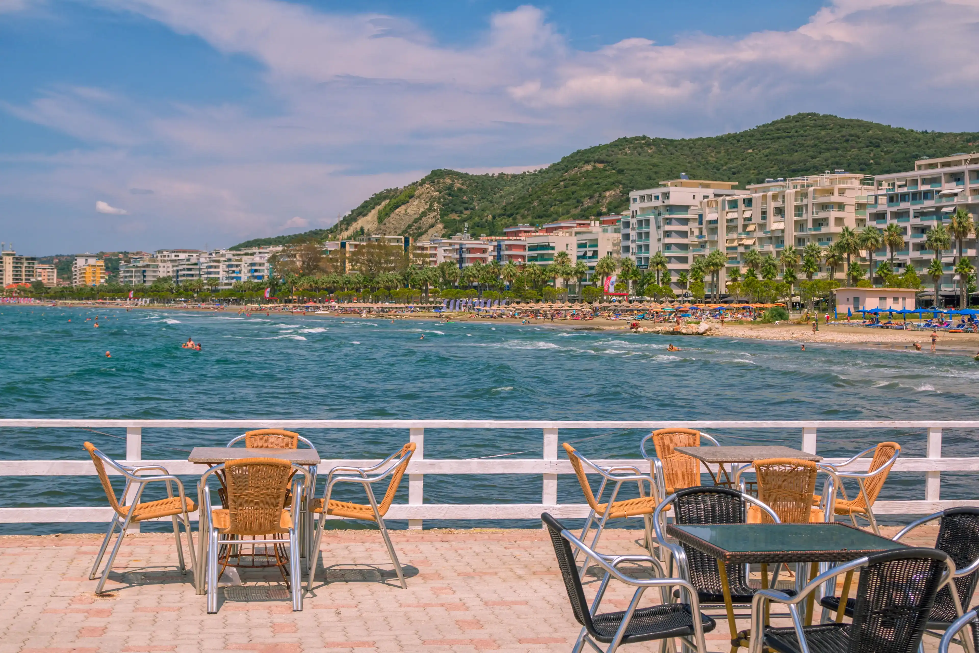 Vlora, Albania - August 7, 2020: summer resort –outdoors café – tables and chairs, turquoise sea water, people swimming, sunbathing on the beach, hotels buildings, blue sky and mountain on background