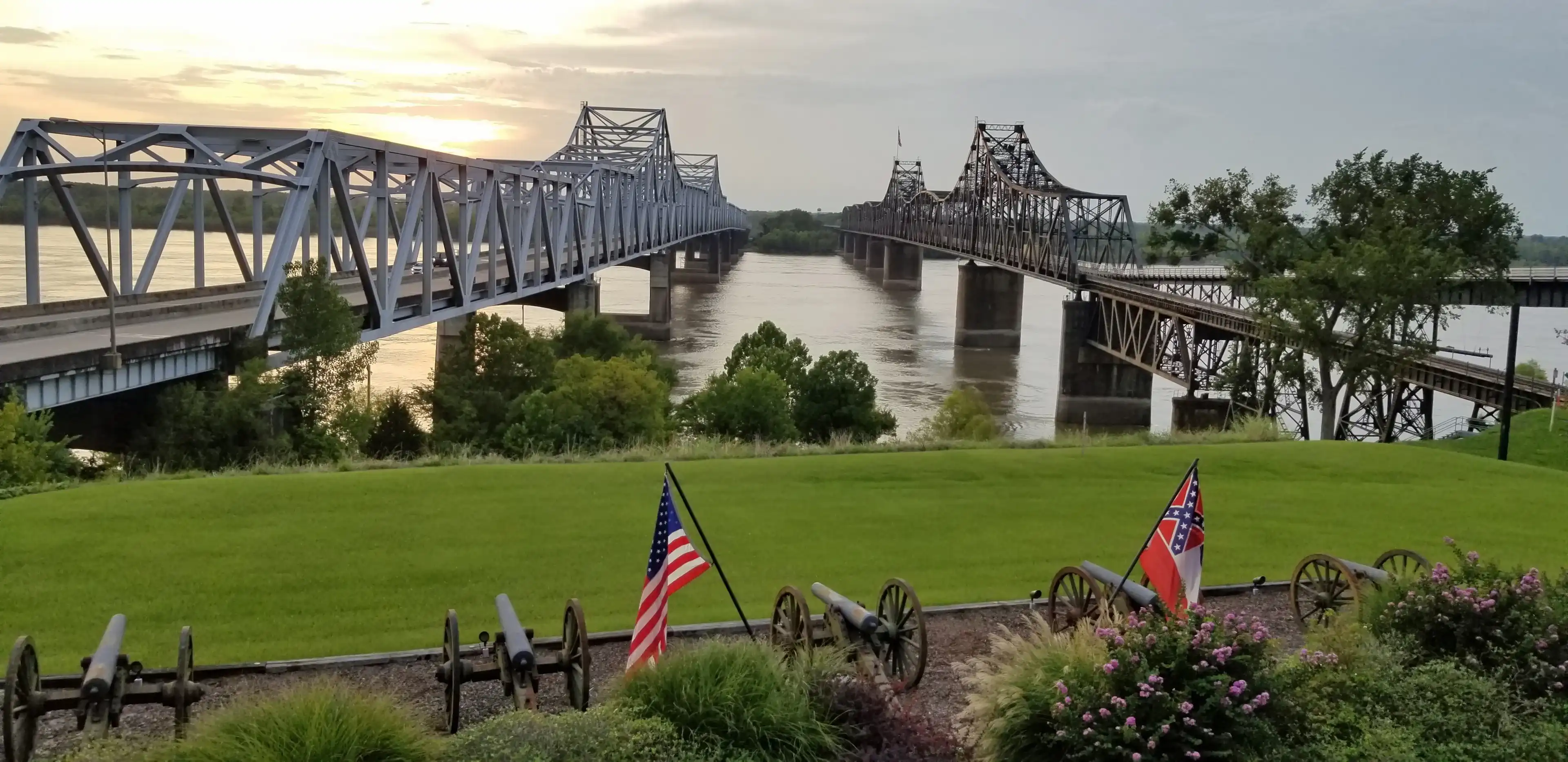 Two bridges crossing the Mississippi River contrasting old and new, railroad and highway, and train and road in Vicksburg, Mississippi with the United States and State of Mississippi flag flying. Two bridges crossing the Mississippi River contrasting old and new, railroad and highway, and train and road in Vicksburg, Mississippi with the United States and State of Mississippi flag flying.