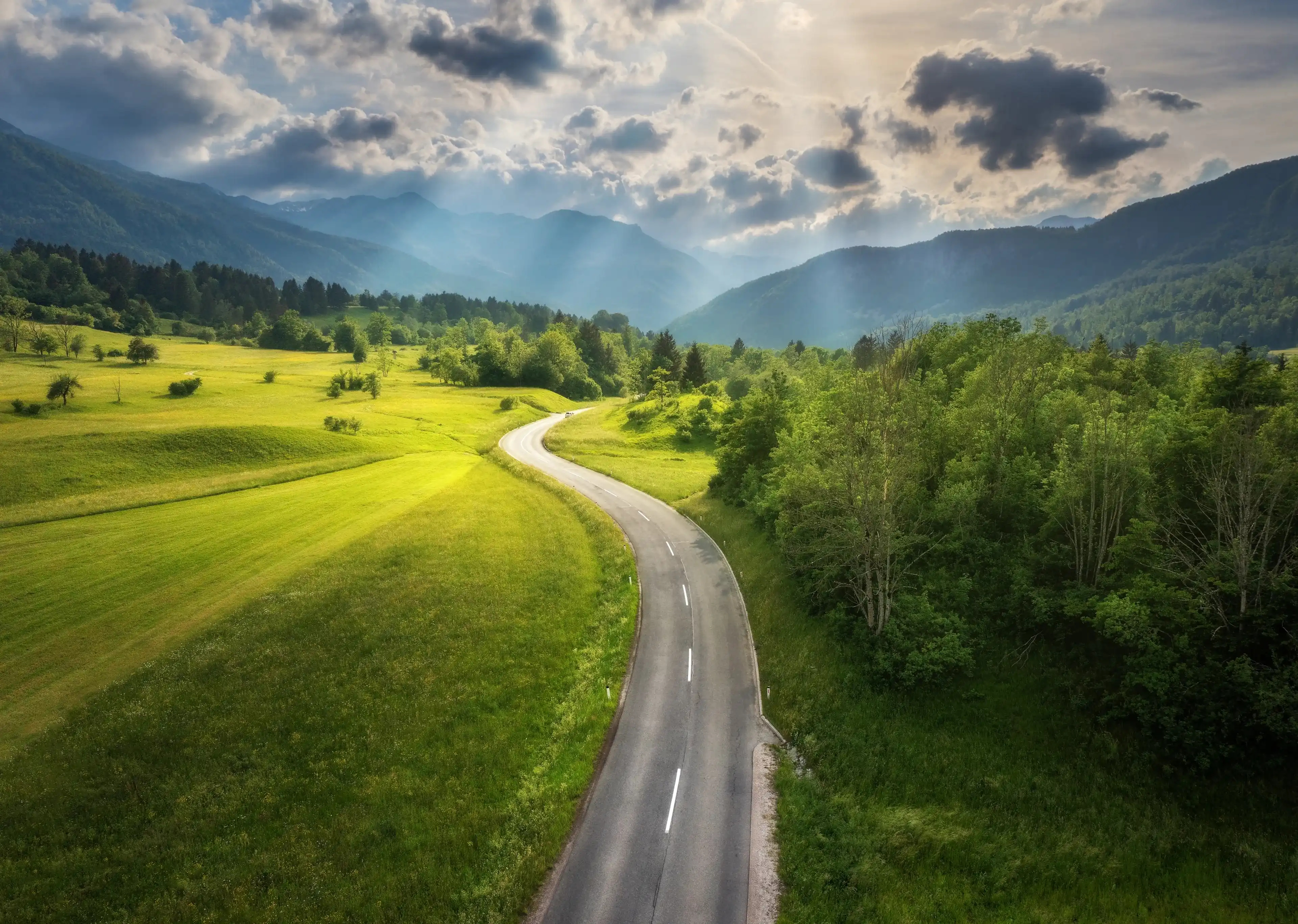 Aerial view of road in green meadows at sunset in summer. Top view from drone of rural road, mountains, forest. Beautiful landscape with roadway, sun rays, trees, hills, green grass, clouds. Slovenia