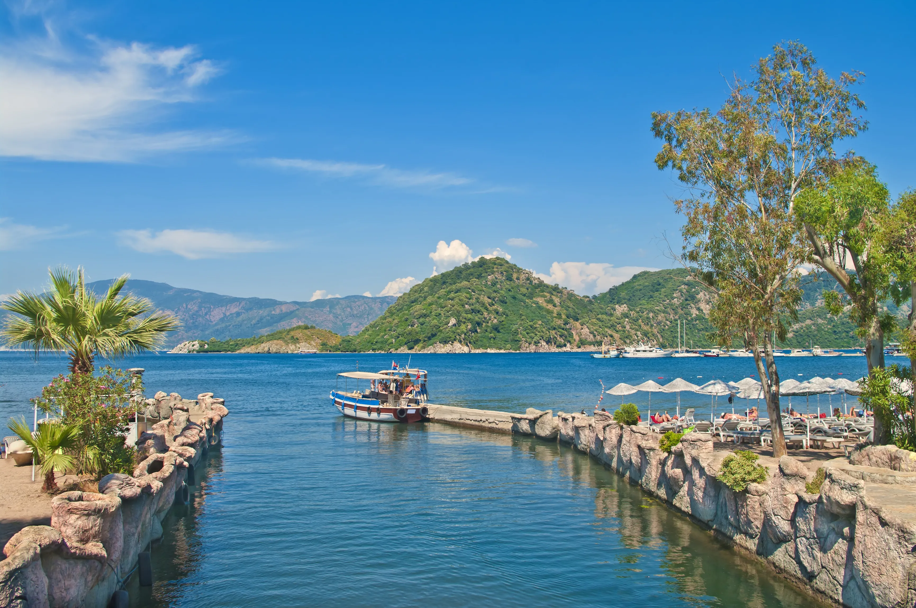 small tourist boat at Icmeler pier in Aegean sea bay with palm trees at foreground and mountains at background, Marmaris, Turkey